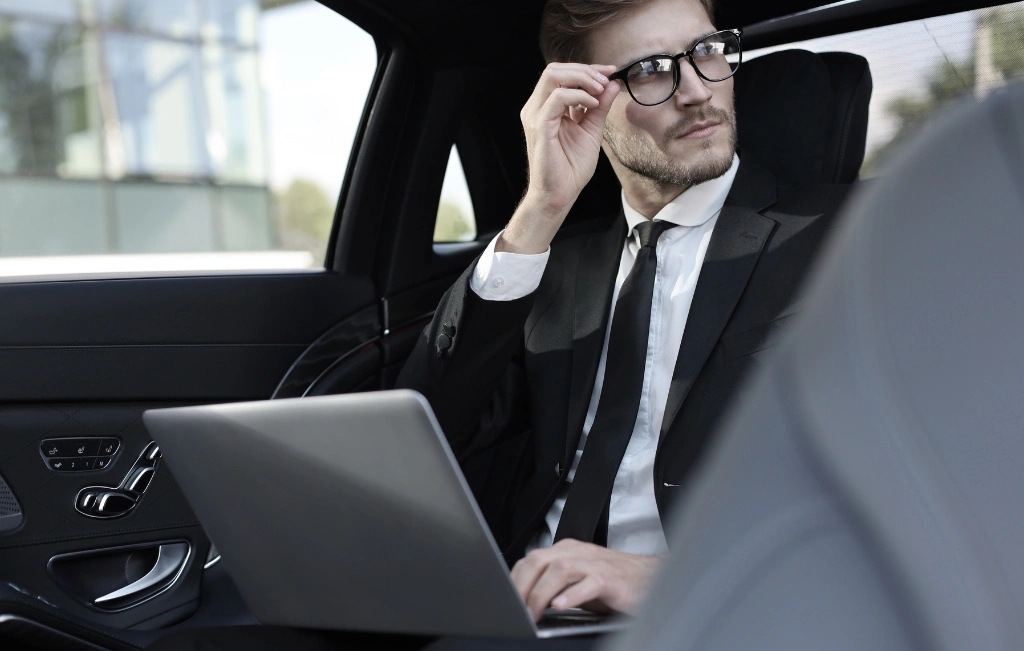 Thoughtful-young-businessman-keeping-hand-on-glasses-while-sitting-in-the-lux-car-and-using-his-laptop-during-airport-car-service